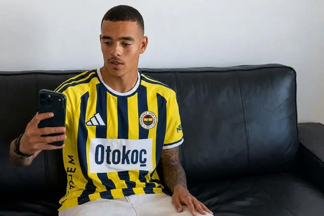 A man in a Fenerbahce jersey sitting on a couch using a smartphone.