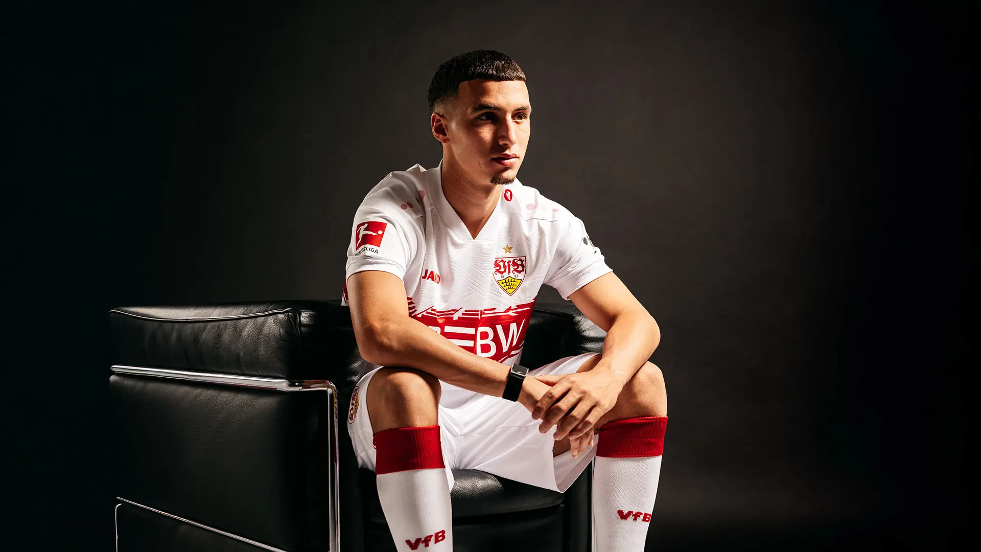 Bilal El Khannouss posing in a white VfB Stuttgart home kit sitting on a black leather chair