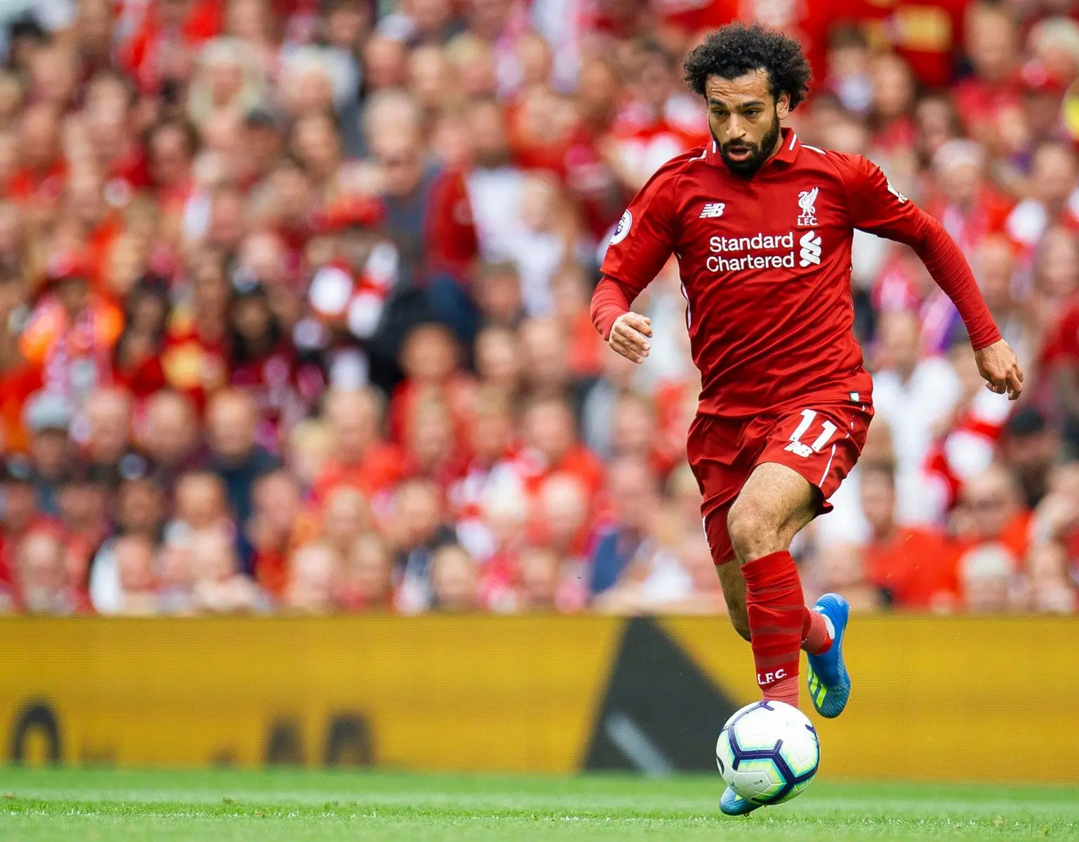Mohamed Salah in a red Liverpool FC kit controlling a football on the pitch with a crowd in the background